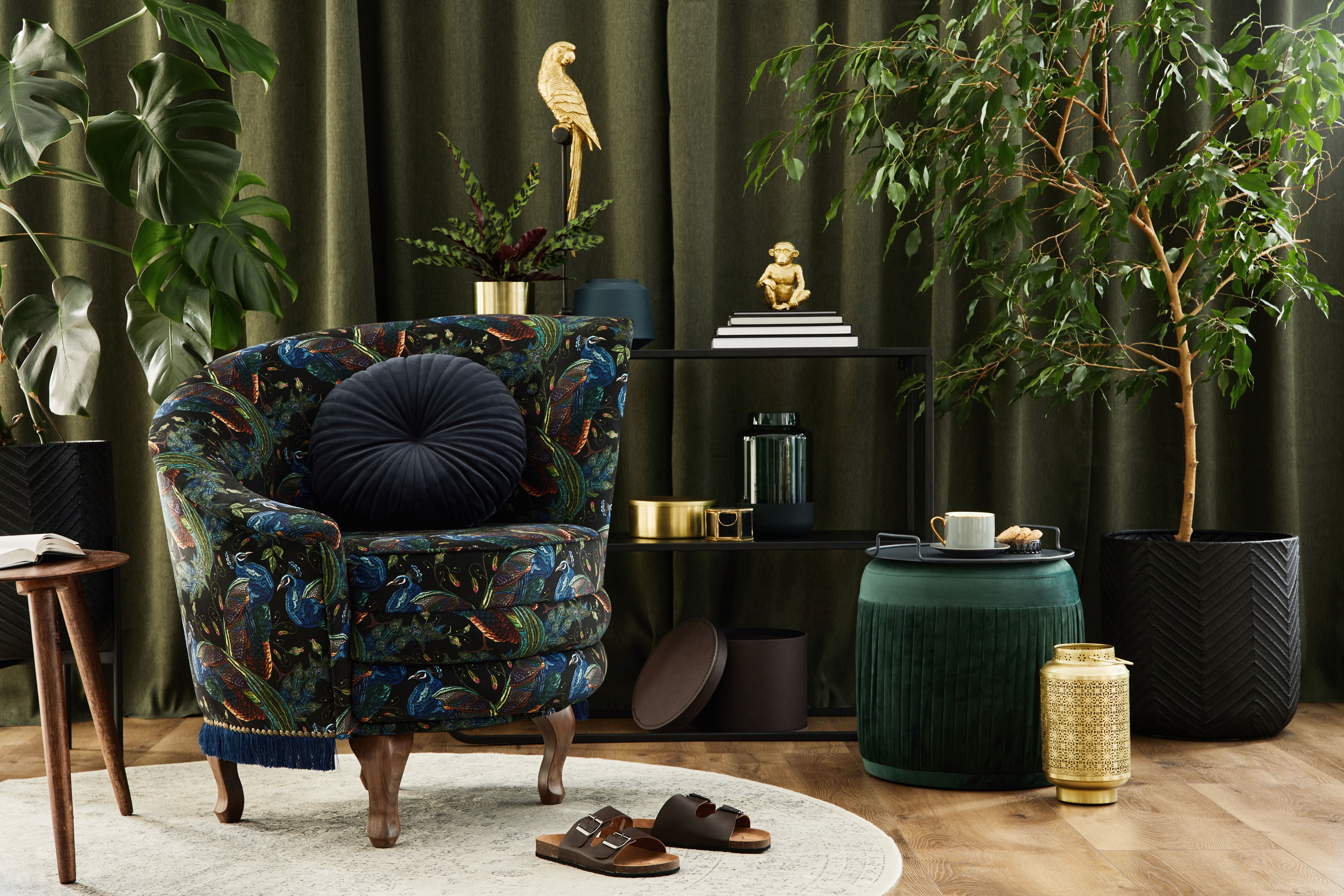 A richly layered corner with deep green velvet drapery, a patterned jewel-toned armchair, brass accent tables, and a grouping of tall potted plants against dark walls.