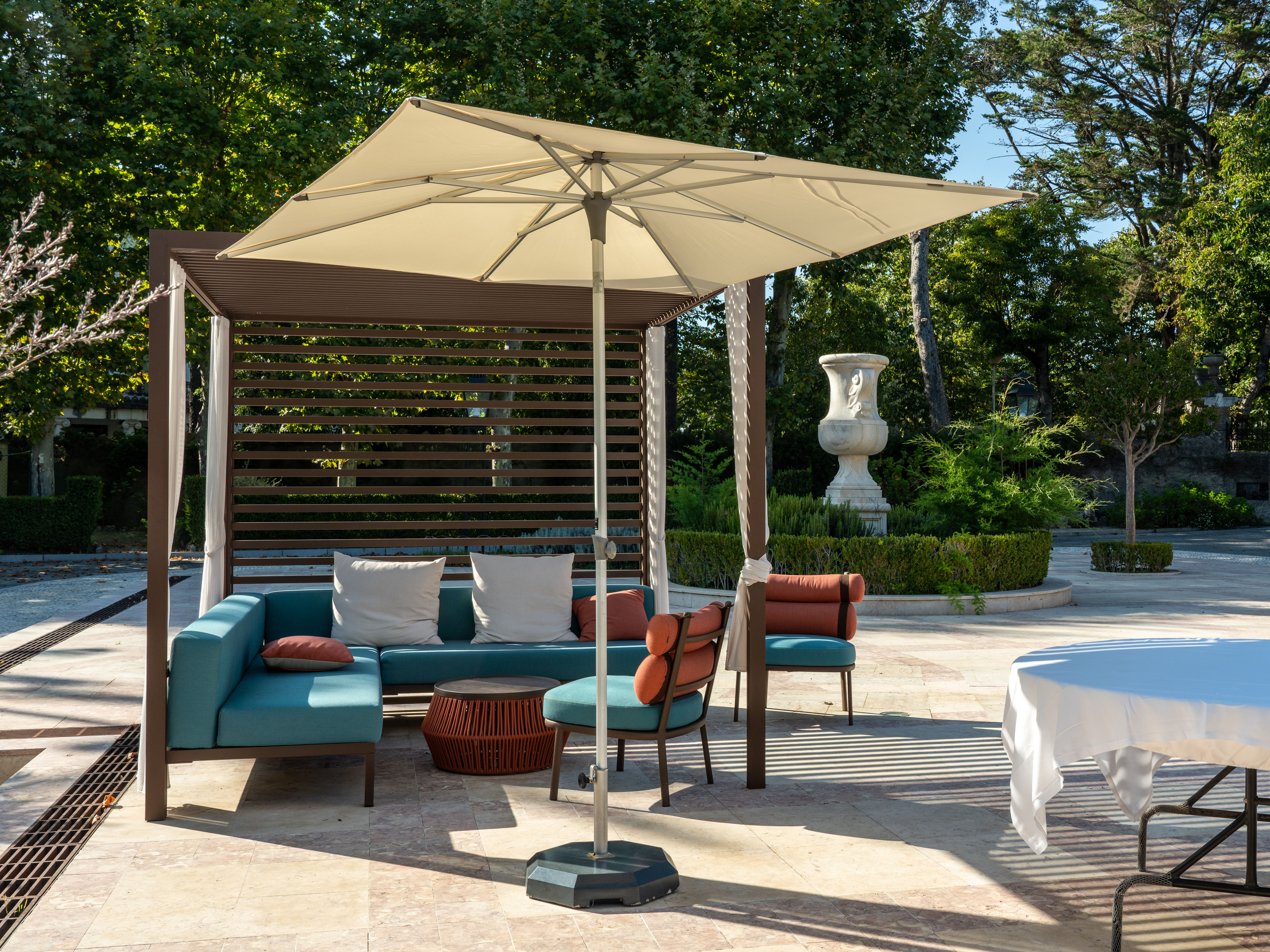 A cantilever umbrella shades a stone-paved patio lounge with teal upholstered sofas and rust-colored throw pillows, a stone urn planter and clipped hedges in the background.