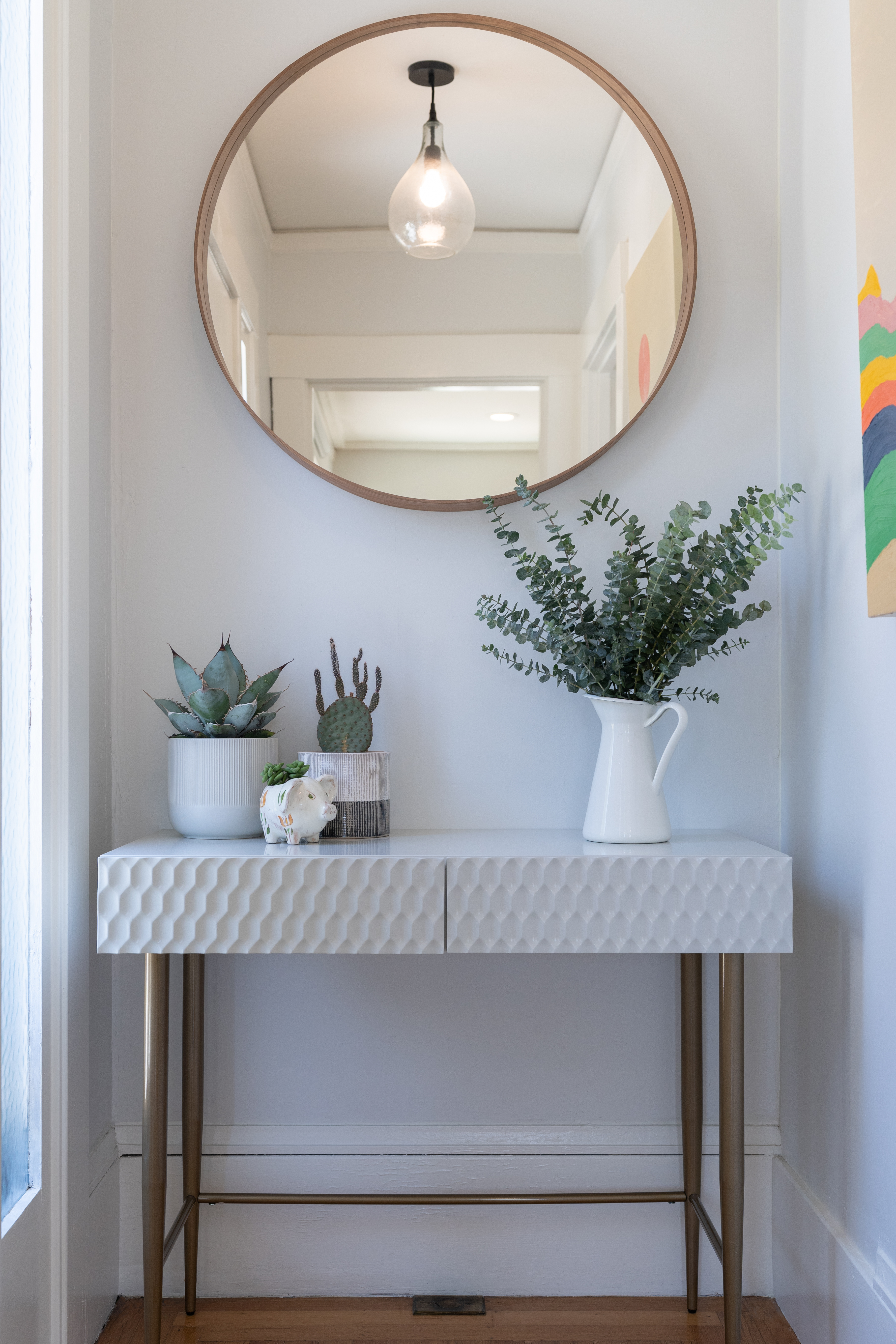 A fluted marble-look console table sits below a round brass-framed mirror, styled with a stoneware vase of eucalyptus and small potted succulents.