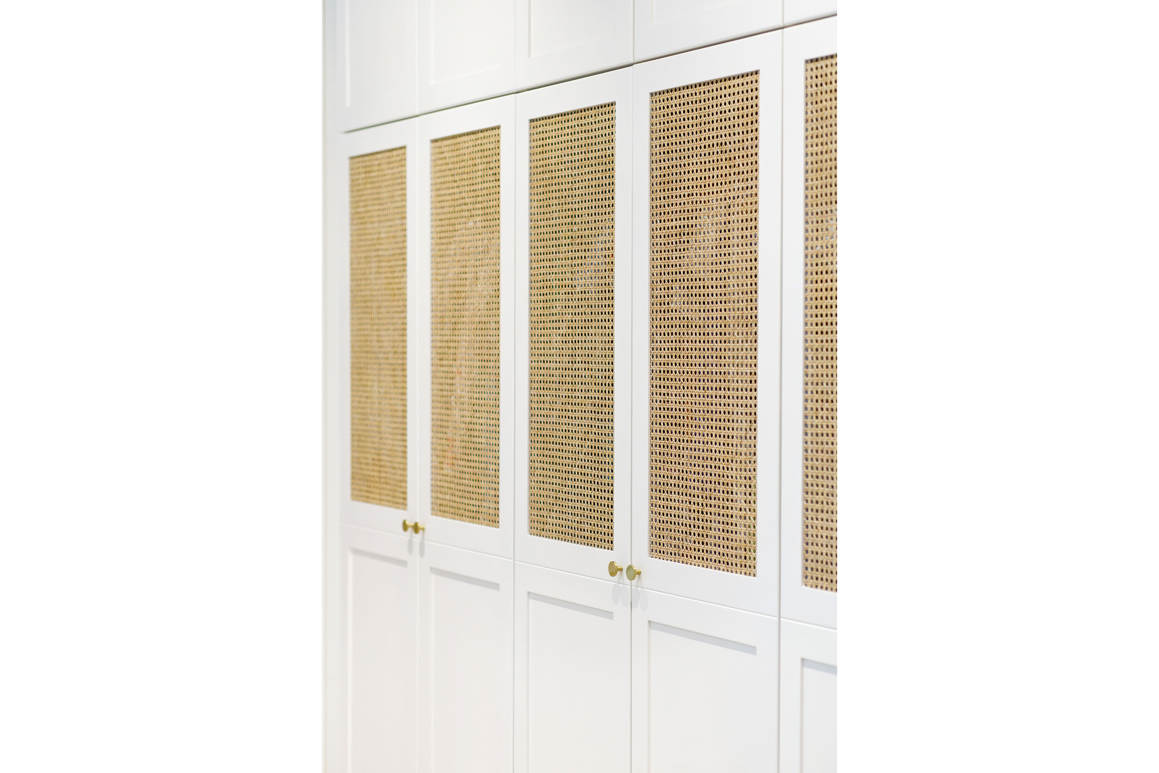 A row of white wardrobe cabinets with woven cane door panels and brass hardware lines a wall.