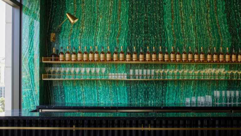 A bookmatched emerald marble back-bar wall holds brass shelves of glassware and bottles.