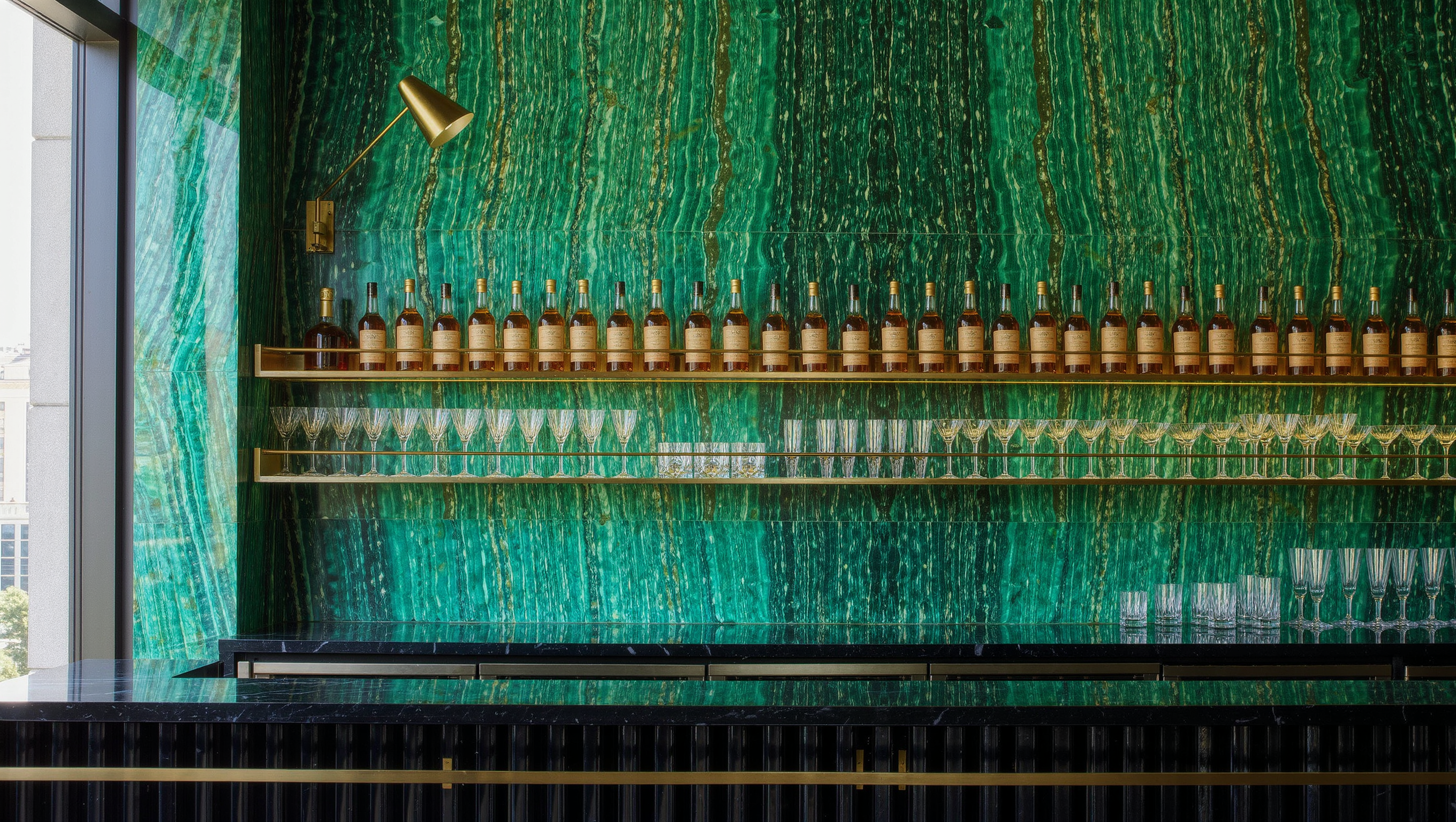 A bookmatched emerald marble back-bar wall holds brass shelves of glassware and bottles.