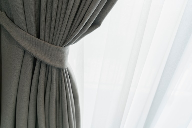 A soft gray-green curtain is tied back beside a sheer inner curtain, with morning light glowing through the window.