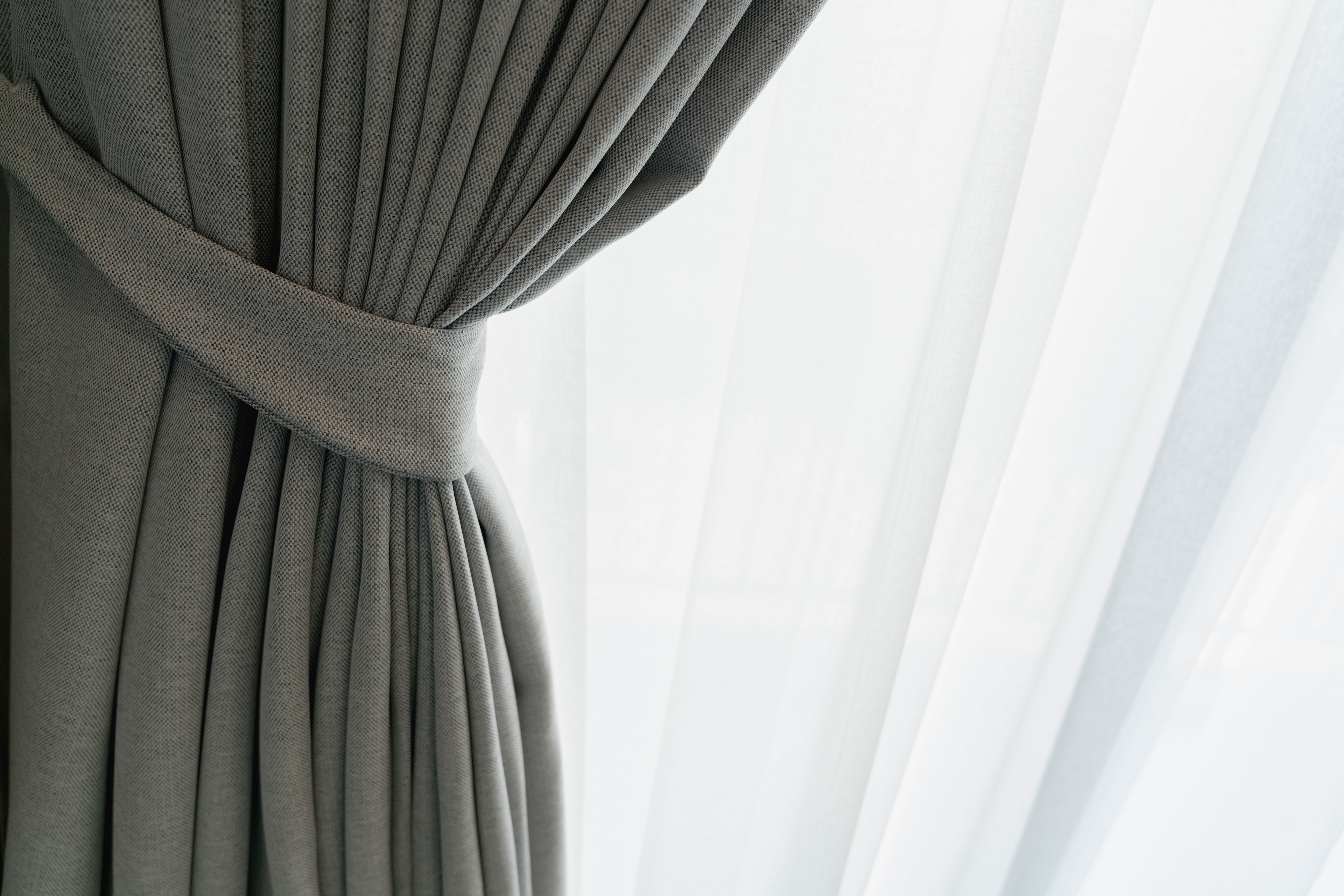 A soft gray-green curtain is tied back beside a sheer inner curtain, with morning light glowing through the window.
