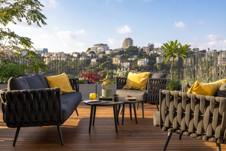 A wooden rooftop deck lined with woven lounge furniture and mustard-yellow pillows overlooks flowering planters and a hillside view.
