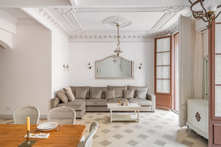 An elegant plaster-molded living room with a chandelier and tiled floor opens through a wide doorway toward the adjoining dining space.