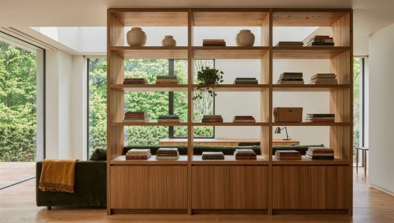A double-sided oak shelving unit dividing a bright room, with a green sofa on one side and a desk zone visible through the shelves.