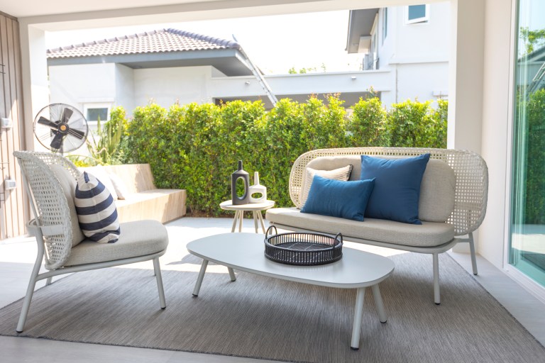 A neutral rattan outdoor seating set with a blue accent pillow is arranged on a home patio backed by a trimmed hedge.
