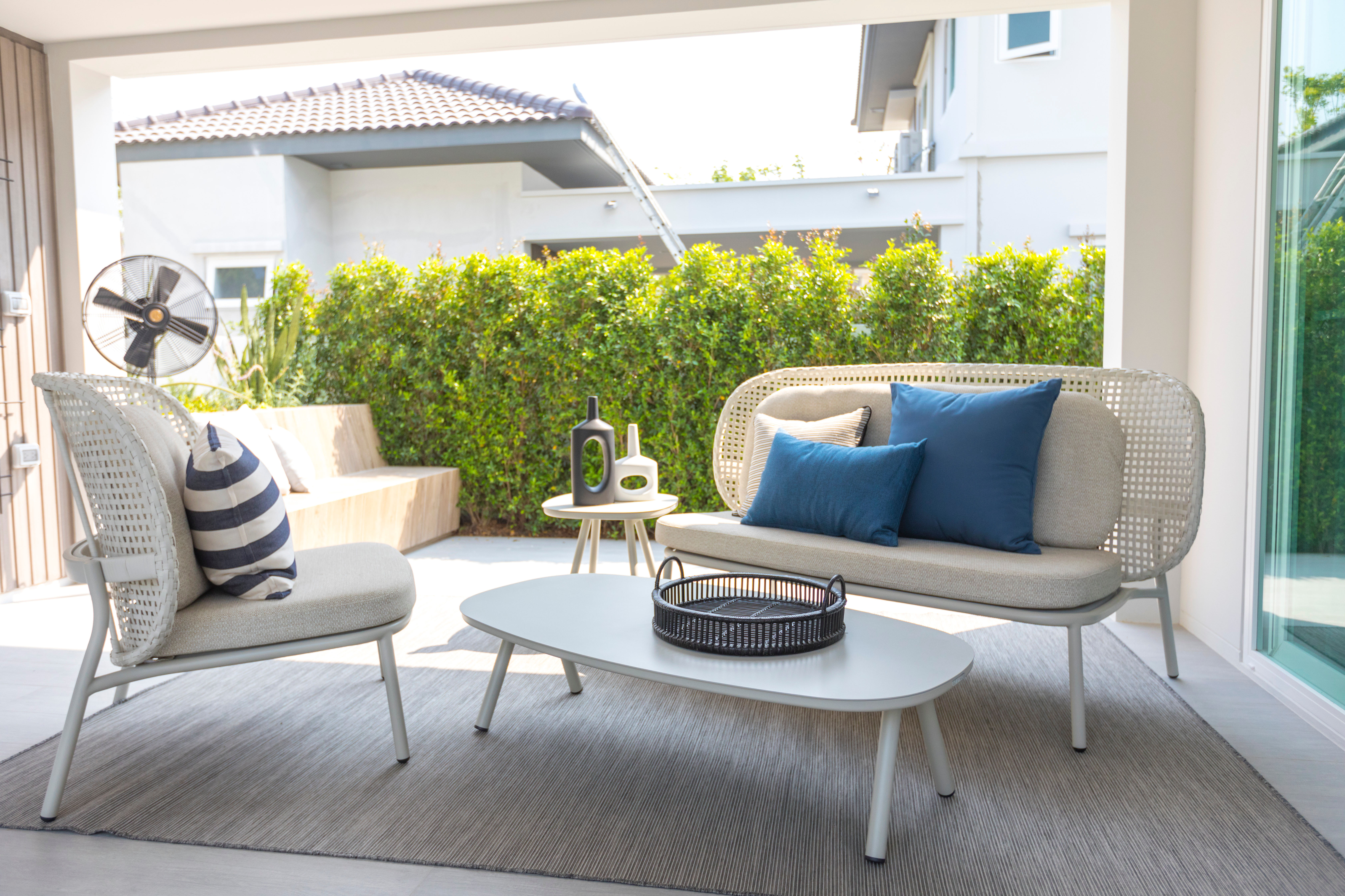 A neutral rattan outdoor seating set with a blue accent pillow is arranged on a home patio backed by a trimmed hedge.