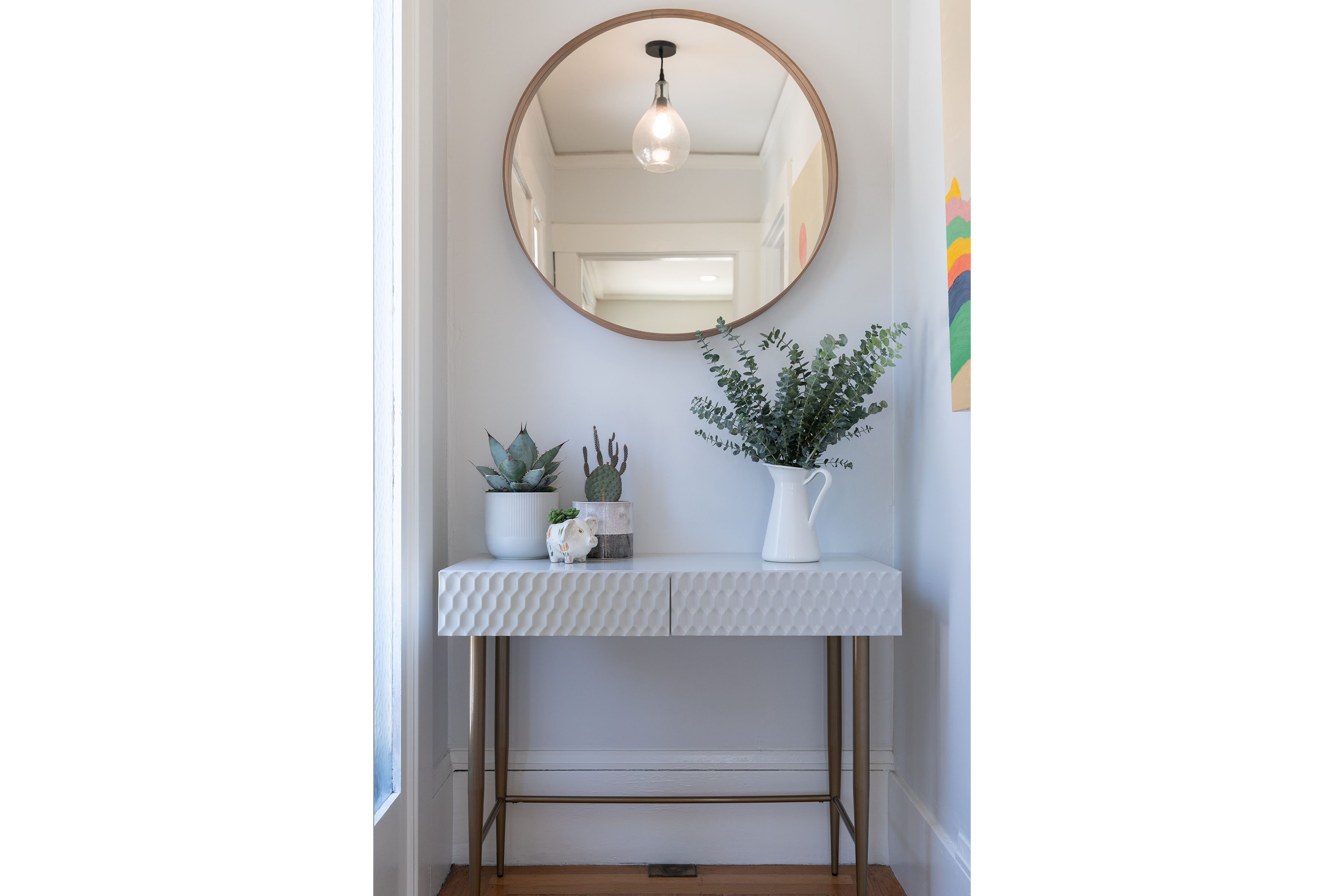 A textured console table topped with potted greenery sits beneath a round brass-framed mirror and a brass pendant light in a bright entryway.