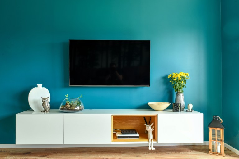 A sleek white low console anchors a teal accent wall beneath a mounted television, styled with a ceramic vase, a glass bowl of flowers, and a brass lantern.