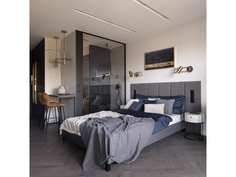 A charcoal-toned bedroom with a tufted headboard and brass wall sconces, divided from the ensuite by a black-framed glass partition.