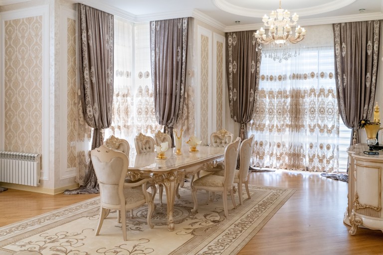An ornate dining salon with a long table and upholstered chairs sits beneath a crystal chandelier, framed by floor-to-ceiling patterned drapery.