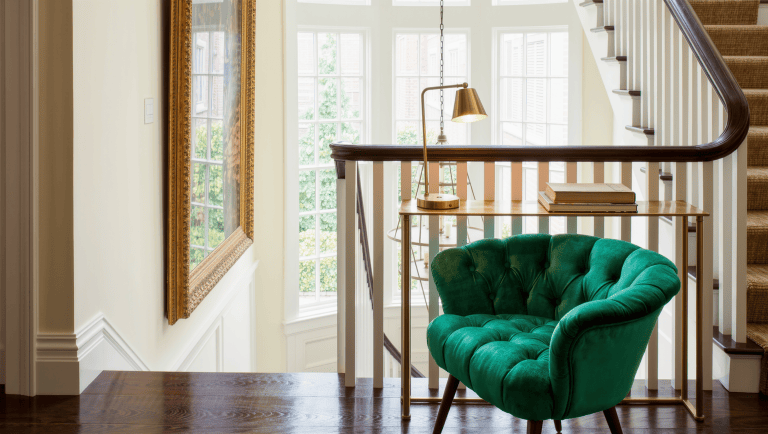 A small velvet chair and brass reading lamp on a stair landing beneath a large gilt-framed artwork.