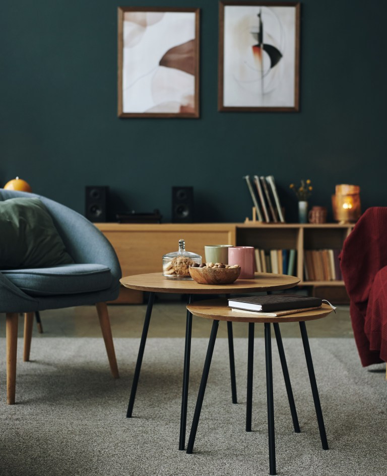 A wood-and-leather bookshelf holding vinyl records sits against a dark forest-green wall, with nesting tables and a blue velvet chair in the foreground.