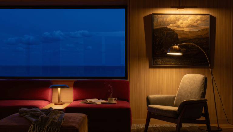 A lounge at dusk with an oxblood sofa lit by low pools of warm light from a table lamp, floor lamp, and picture light.