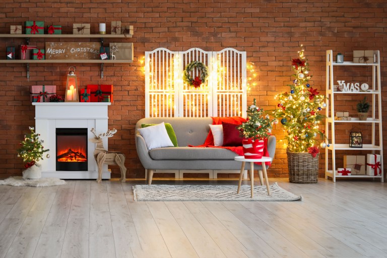 An open shelving unit beside a decorated tree holds mixed ornaments, wrapped gifts, and warm string lights against an exposed brick wall.