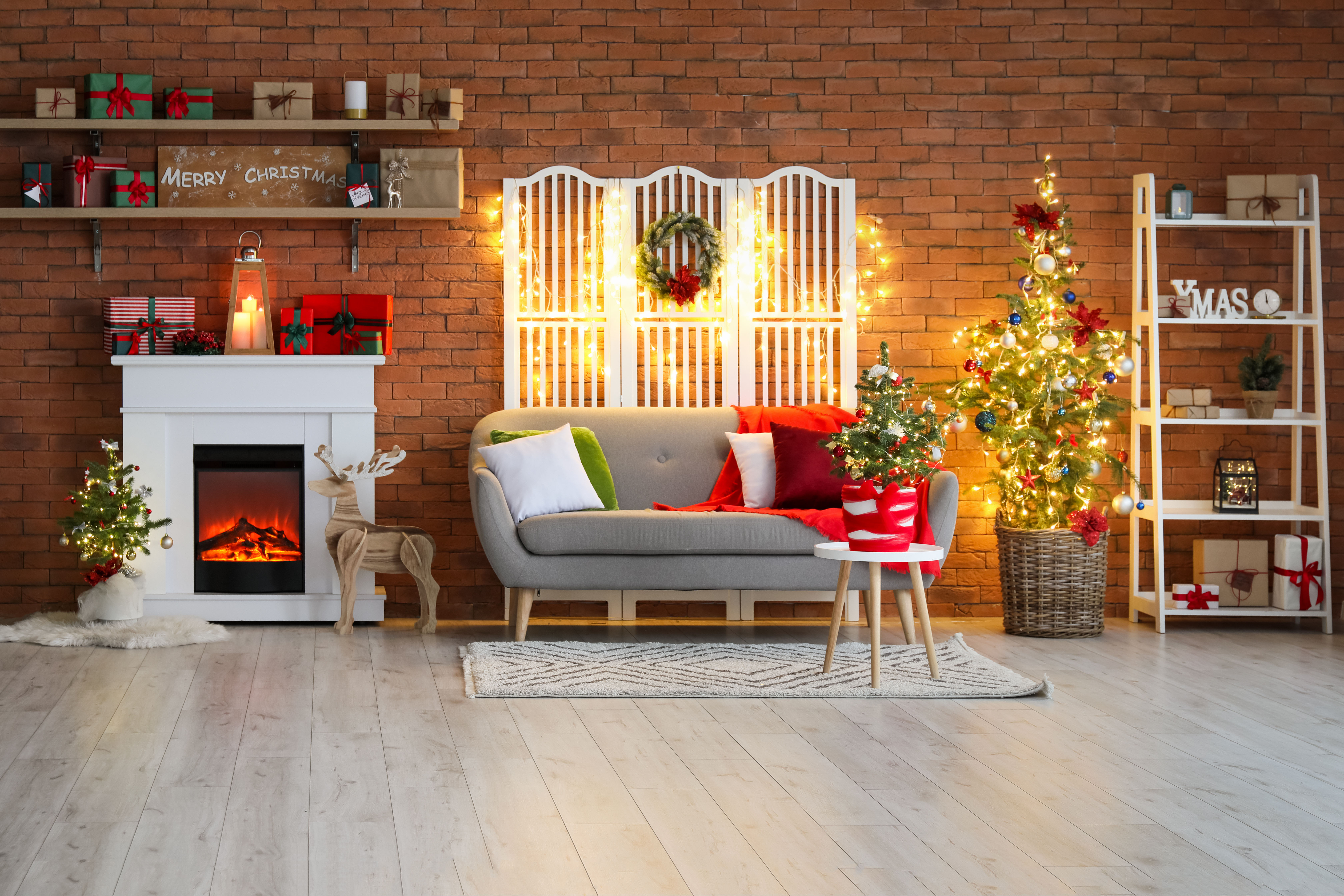 An open shelving unit beside a decorated tree holds mixed ornaments, wrapped gifts, and warm string lights against an exposed brick wall.