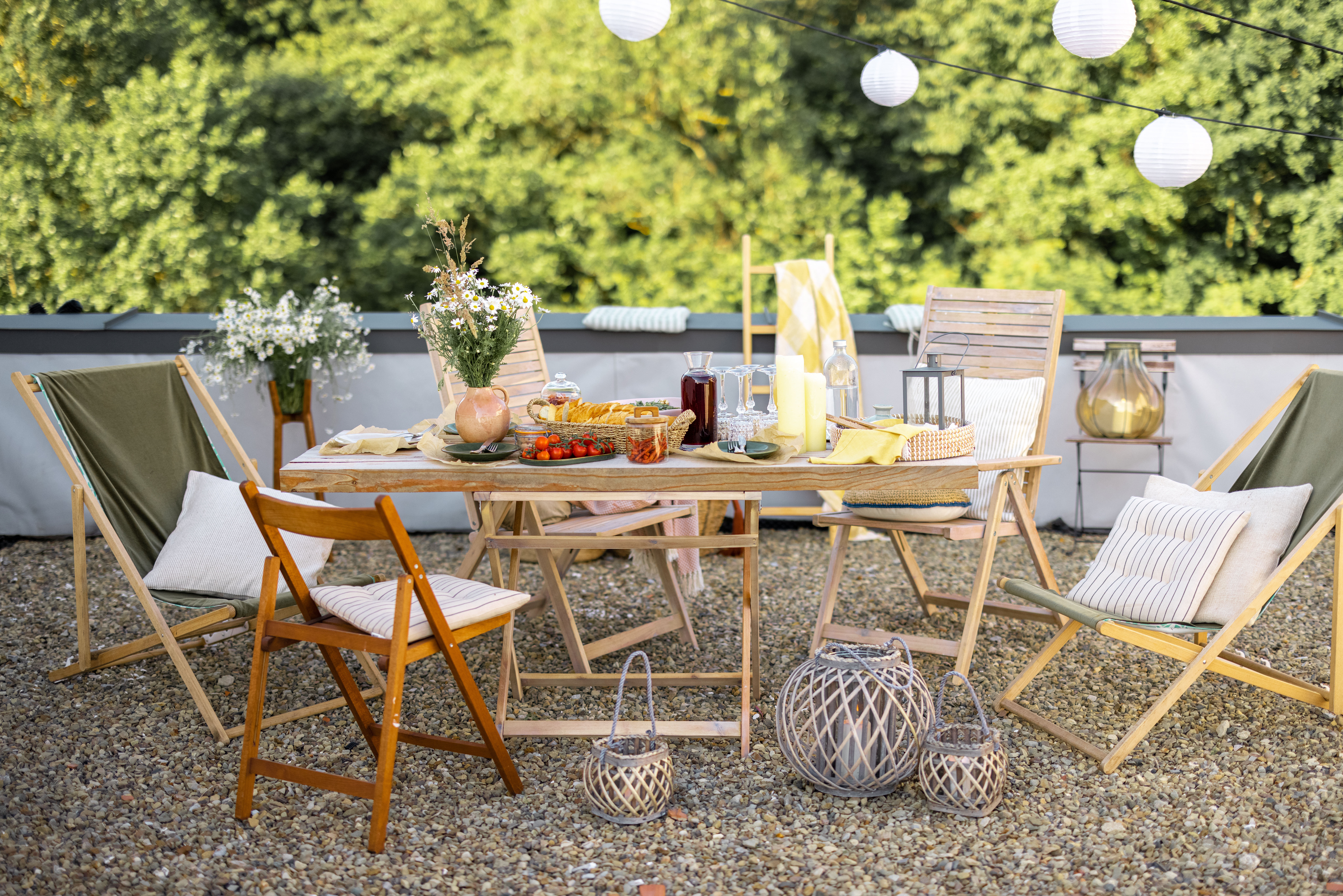 A long wooden table set for a small outdoor supper on a gravel terrace, dressed with folding chairs, paper lanterns, and greenery along the boundary.