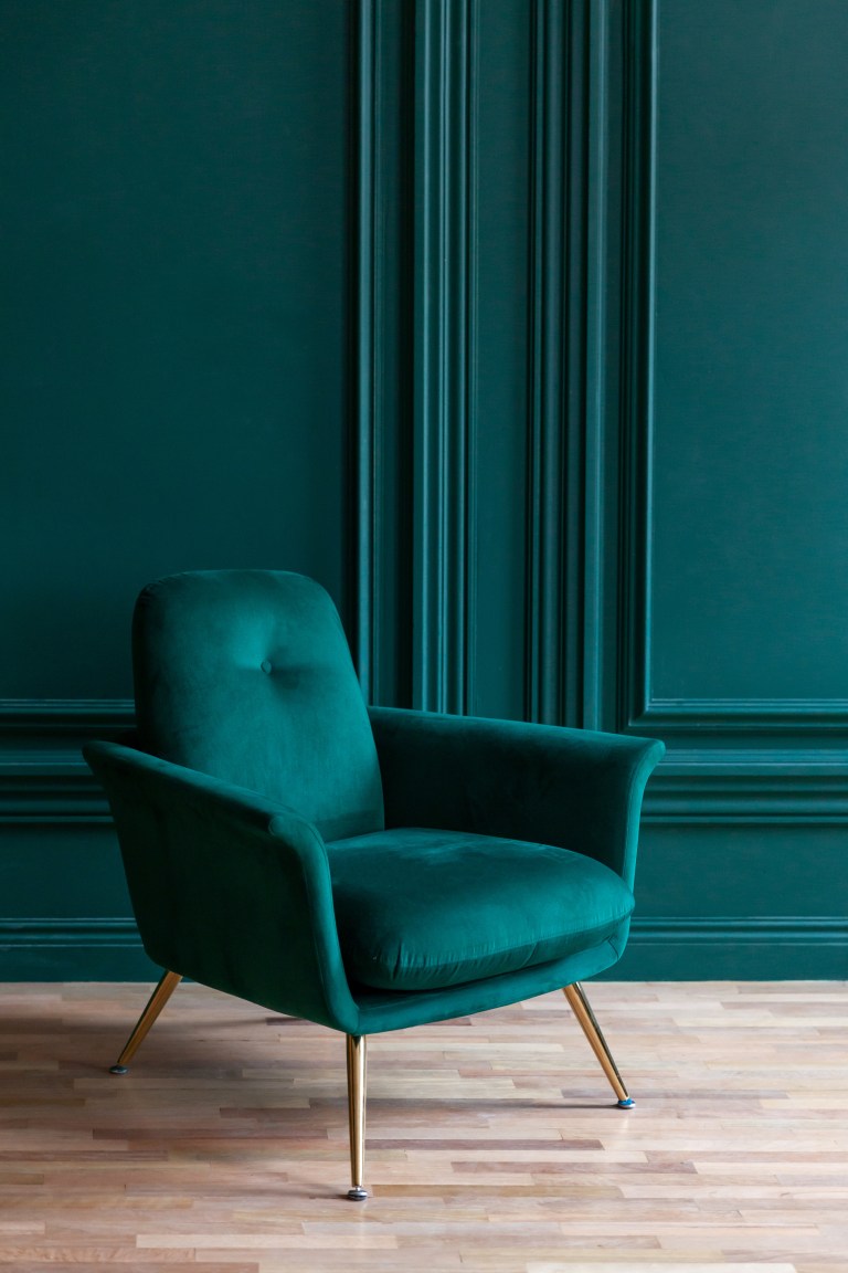A single deep emerald velvet armchair with slim brass legs stands against a matching green paneled wall.