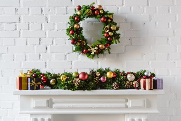 A white mantel is layered with a garland of baubles, wrapped gifts, and a matching wreath in red, gold, and green above it.