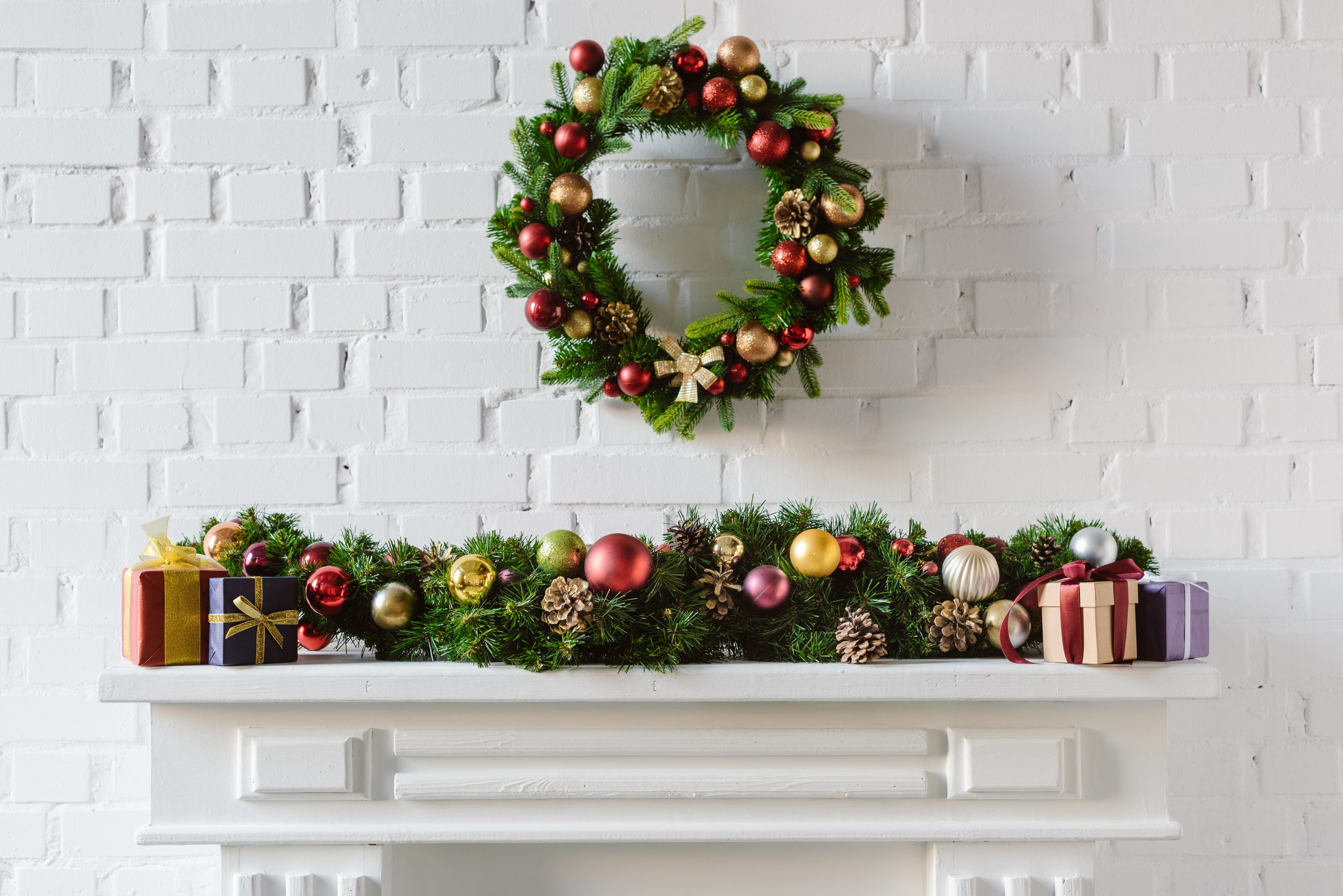 A white mantel is layered with a garland of baubles, wrapped gifts, and a matching wreath in red, gold, and green above it.