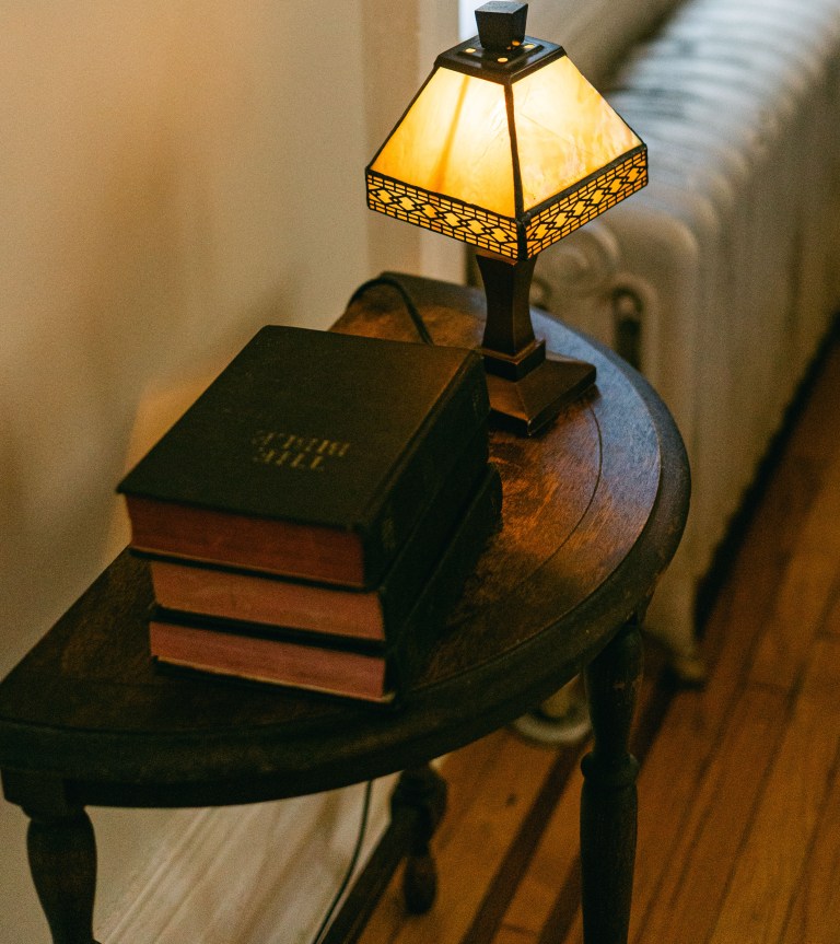 A round wooden side table holds a stack of leather-bound books beneath a lit stained-glass lamp, set beside a radiator and warm wood floor, ready to welcome an arriving guest.