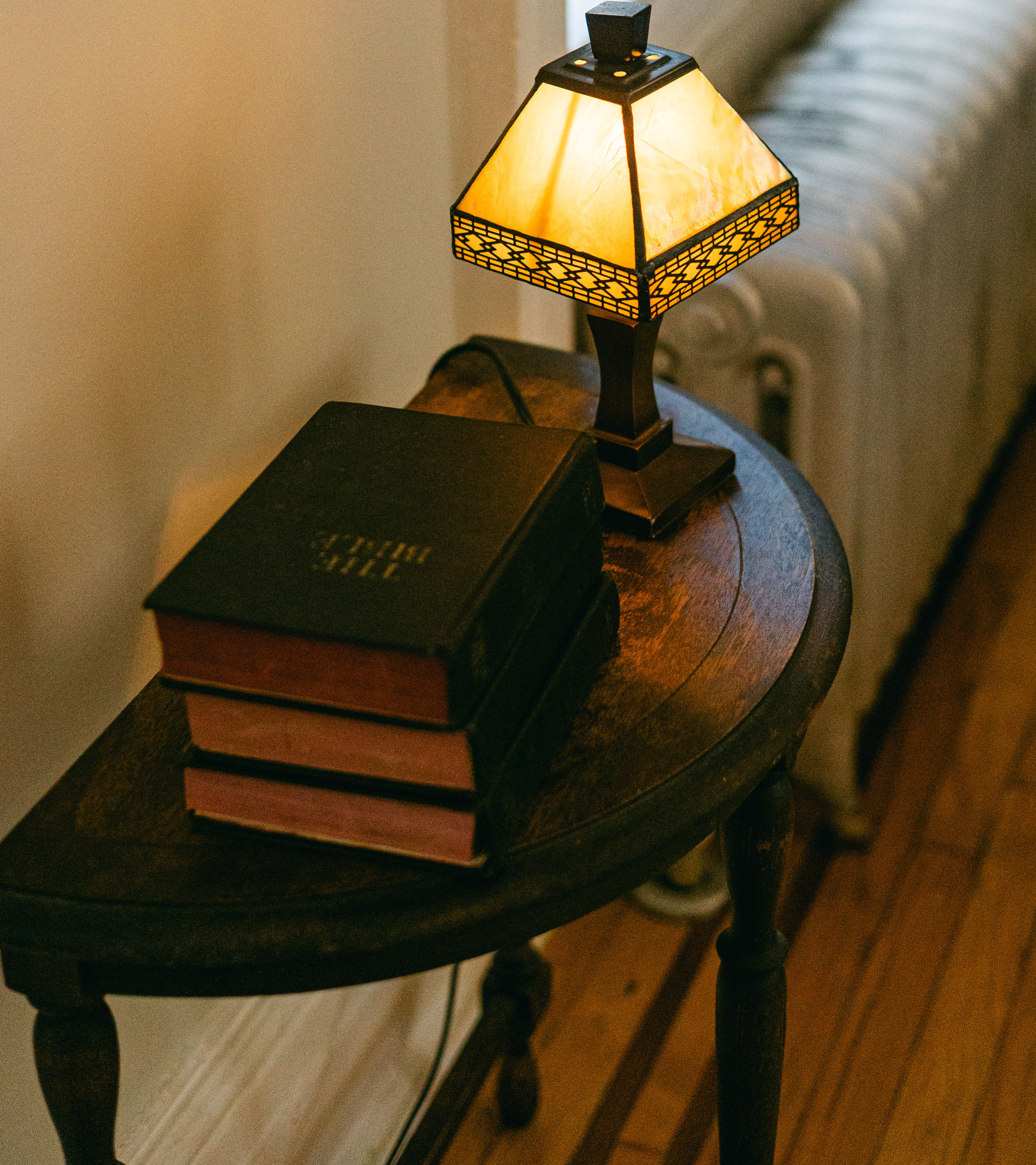 A round wooden side table holds a stack of leather-bound books beneath a lit stained-glass lamp, set beside a radiator and warm wood floor, ready to welcome an arriving guest.