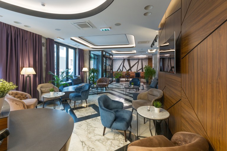 A warm hotel-style lounge with teal velvet armchairs, brass-legged side tables, and rich wood paneling arranged along the room's outer edge.