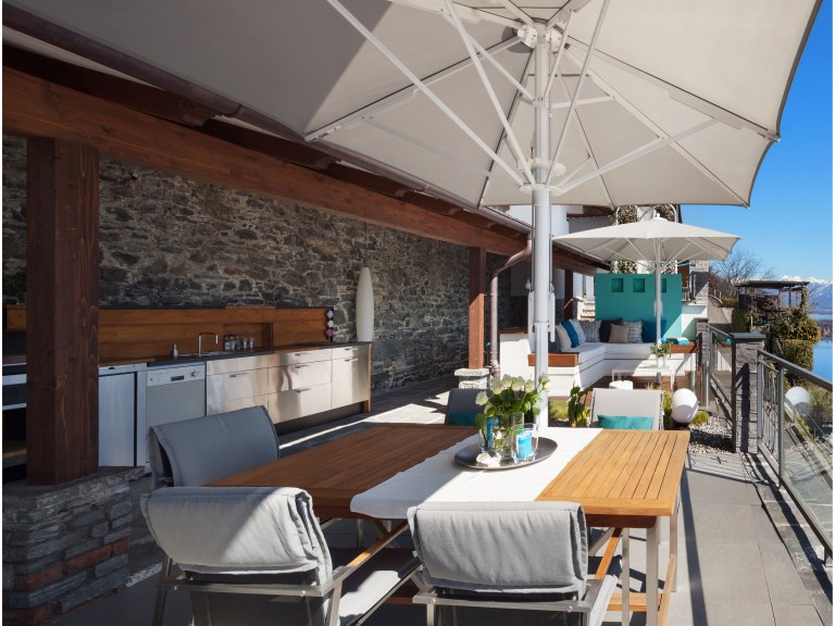 A stone-walled rooftop terrace shaded by a large market umbrella, with a teak dining table set beneath it and an outdoor kitchen counter along the wall, sea view beyond the railing.