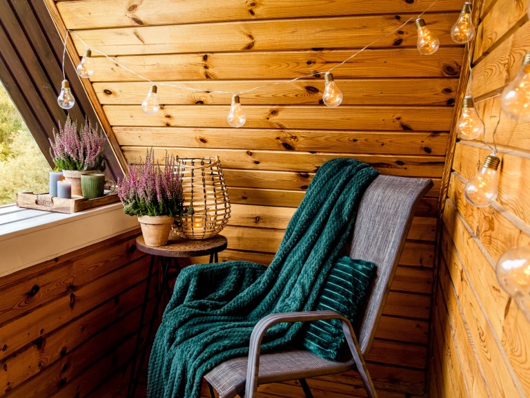 A knotty-pine sheltered balcony corner strung with warm lights, holding a wicker chair draped in a deep teal throw beside potted heather.