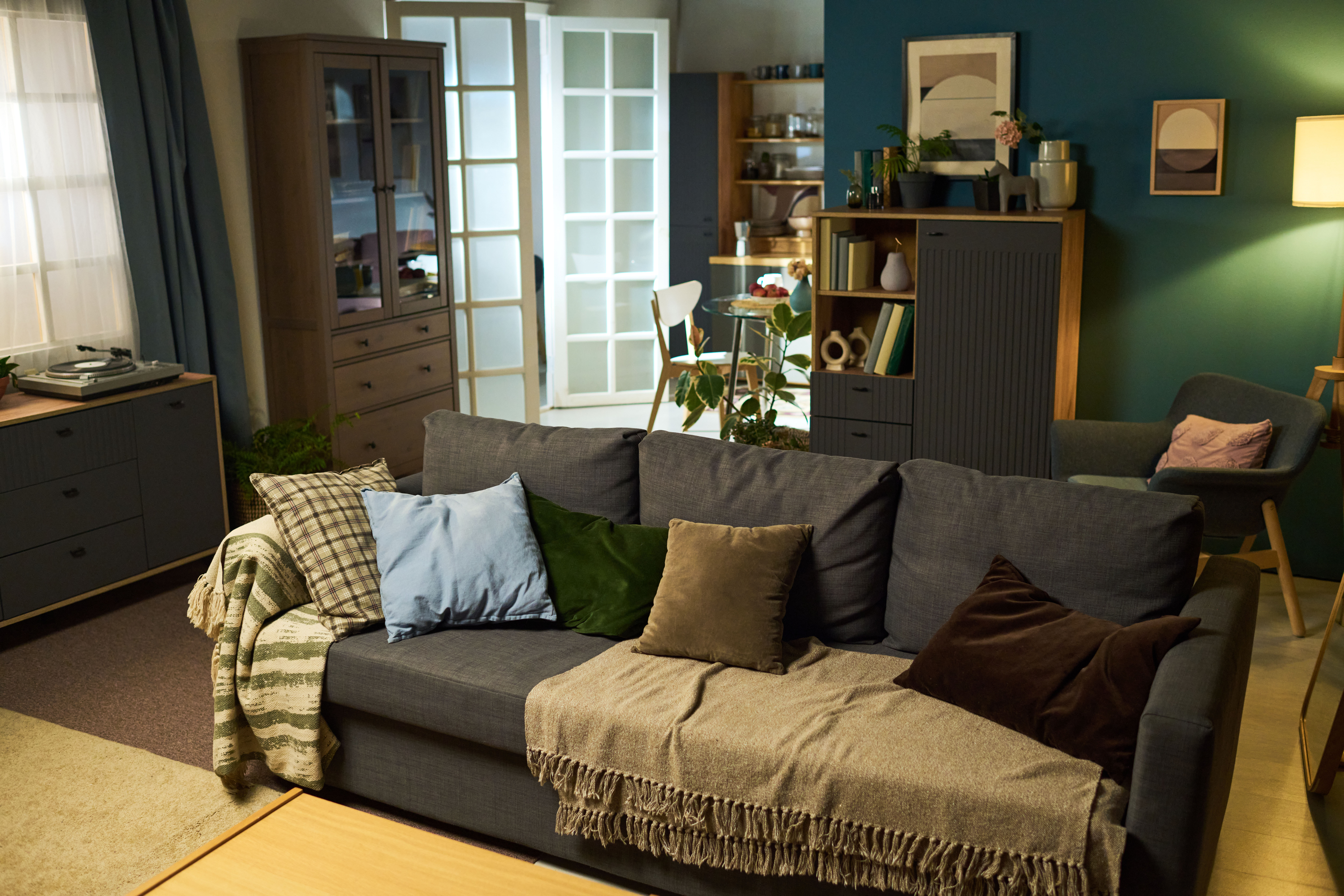 A deep-gray sectional sofa with rust and patterned pillows sits back-to-back with a dining area, its dark teal wall and built-in shelving marking a clear zone change.