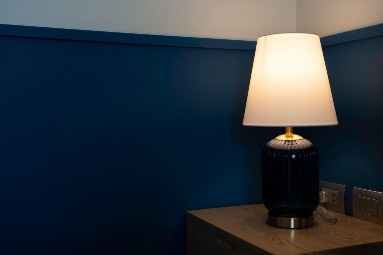 A tall table lamp with a white drum shade and a brass-ringed black base glows warmly in the corner of a deep navy blue room.