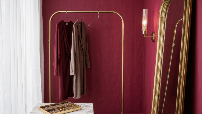 A brass valet rail with two draped silk garments beside a marble-top bench and a gilt-framed mirror.