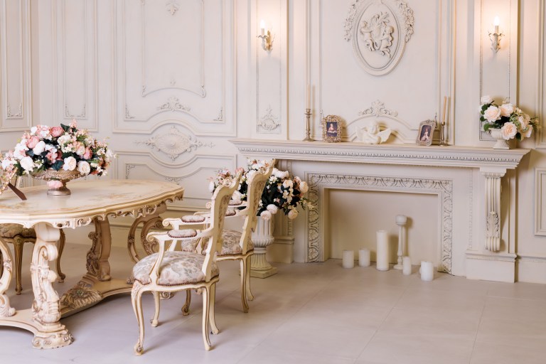 A gilded console table and carved chair stand before an ornate fireplace mantle in a classic cream-and-gold living room, forming the room's first arrival view.