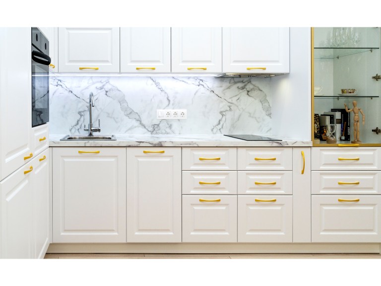 A row of white shaker cabinets with brass hardware surrounds a marble backsplash, keeping cookware and dishware tucked behind matching doors.