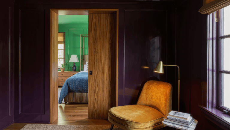 Small aubergine-paneled room with a walnut pocket door half-recessed into the wall and a mustard velvet chair in the space a door swing would claim.