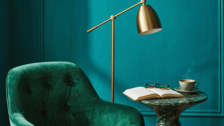 Green marble side table beside an emerald velvet armchair holding an open book, reading glasses, and a steaming cup, against a teal wall.