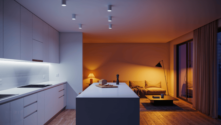 An open-plan space at dusk with the kitchen in crisp neutral-white task light and the lounge glowing amber from dimmed lamps.