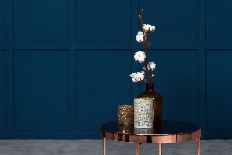 A deep cobalt paneled wall behind a round rose-gold side table styled with a dark vase of white cotton branches.