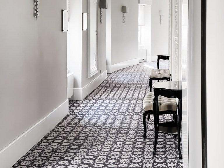 A patterned tile runner leads down an elegant hallway lined with tall doorways, wall sconces, and a pair of black bistro chairs.