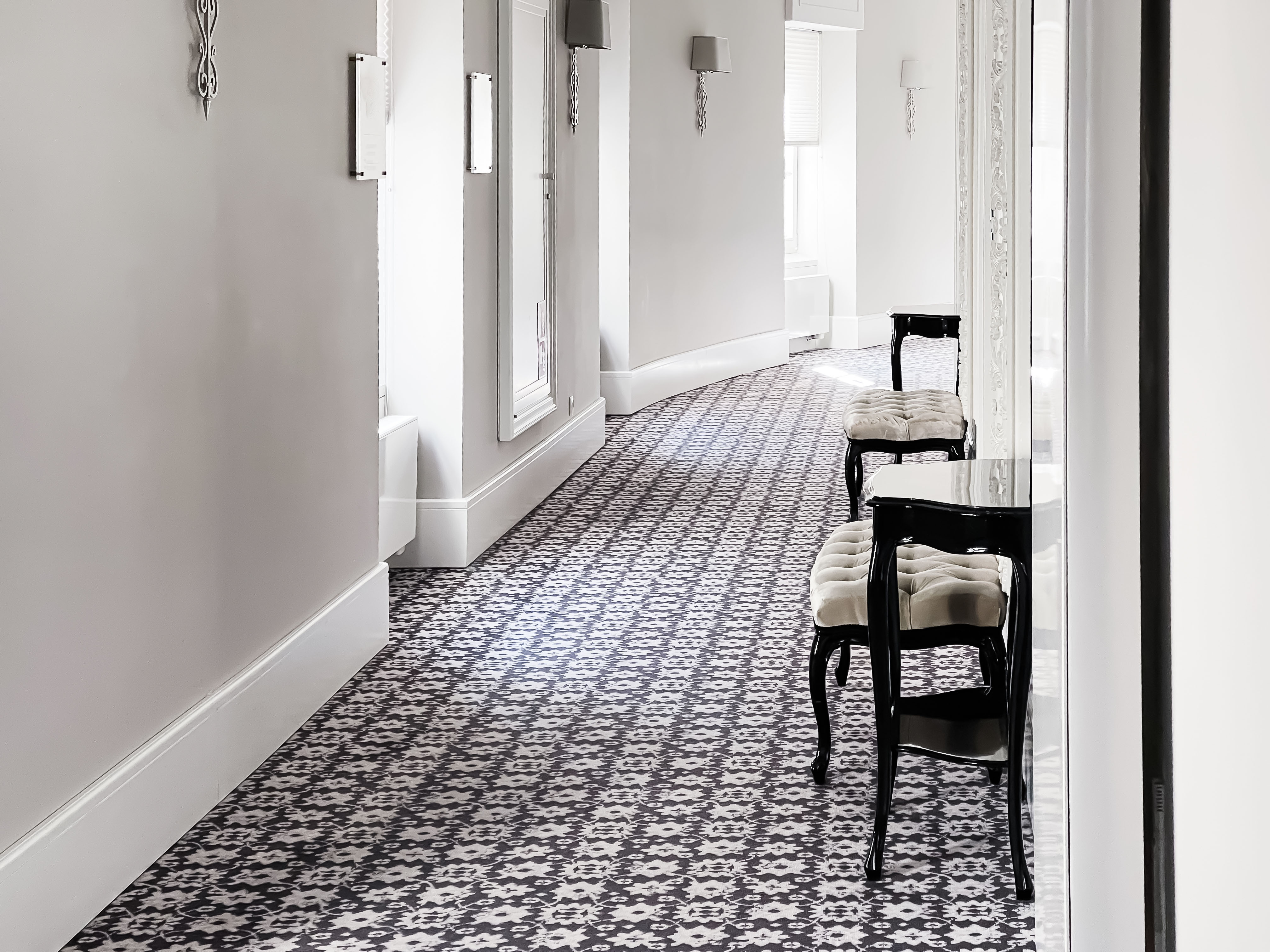 A patterned tile runner leads down an elegant hallway lined with tall doorways, wall sconces, and a pair of black bistro chairs.