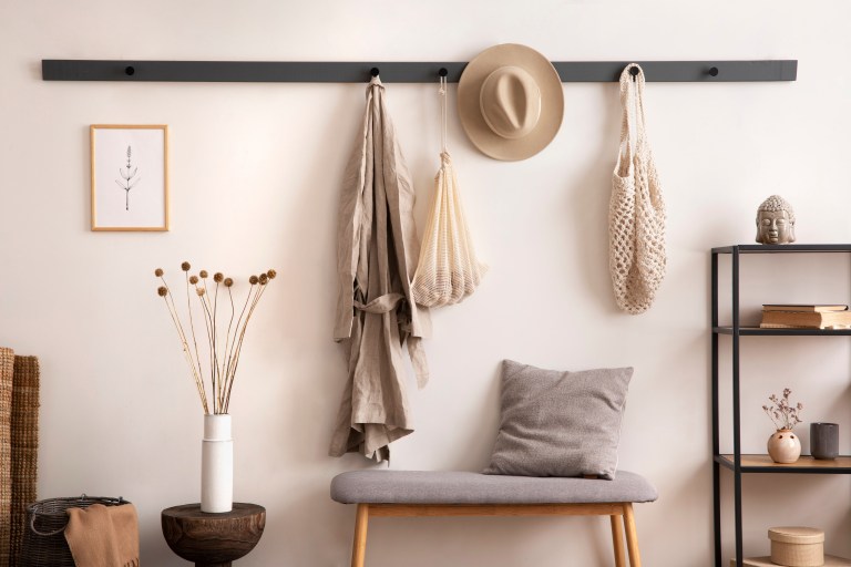 A black wall-mounted rail fitted with hooks holds a hat, tote bags, and a scarf above a cushioned bench, keeping the entryway floor clear.