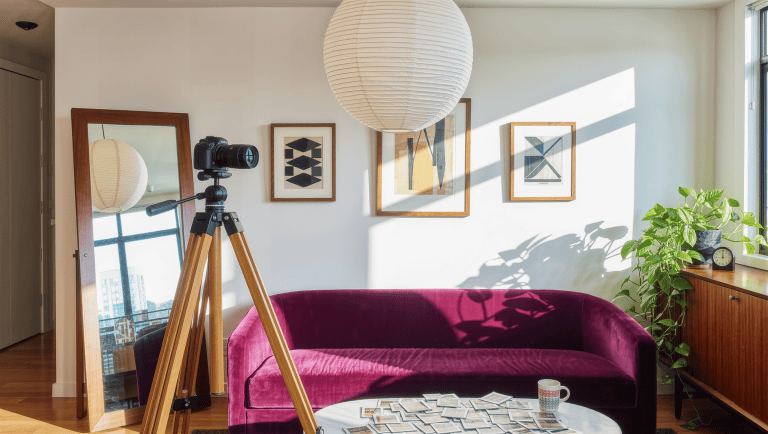 A camera on a wooden tripod aimed at a composed living room with a plum velvet sofa, leaning floor mirror, and framed abstract prints.