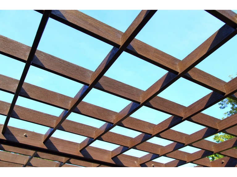 A close view of a wood pergola's crossed beams framing an open sky, warm-toned timber catching the light.