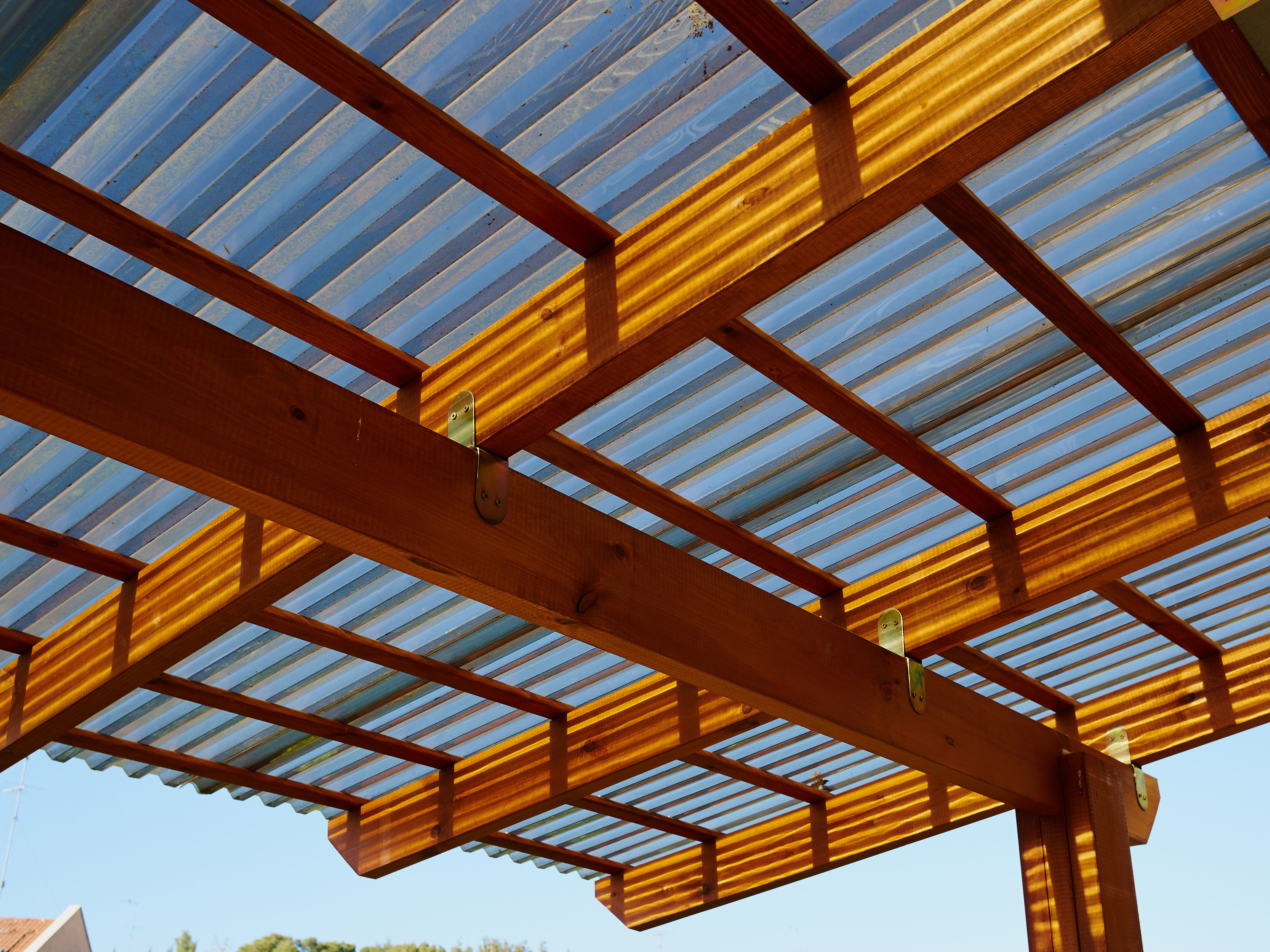 Sunlight filters through the slatted wood beams and polycarbonate roof of a modern pergola against a clear blue sky.