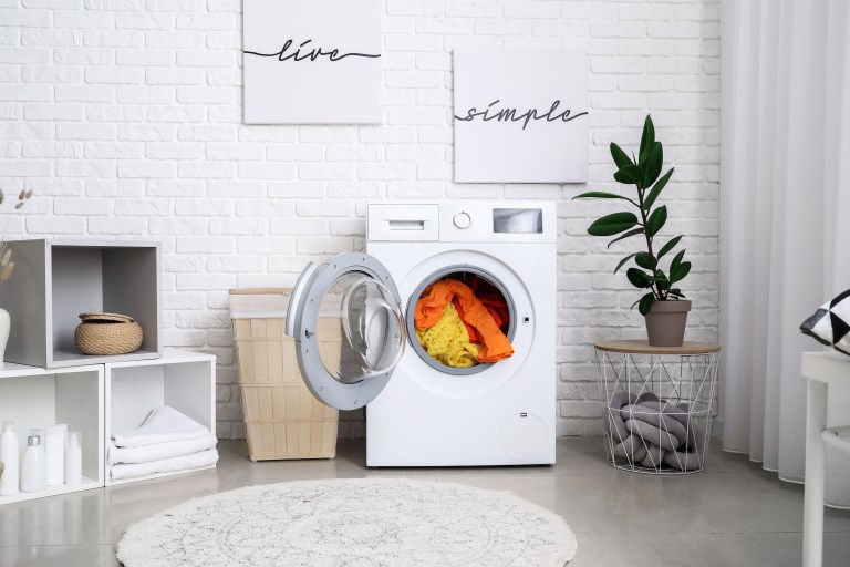A woven laundry basket and stacked baskets sit beside a front-loading washer in a bright white laundry room with a round jute rug marking the walkway.